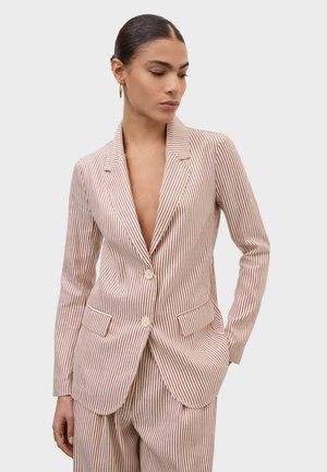 Femme portant un costume crème à fines rayures verticales rouges, boucles d'oreilles créoles dorées et bagues, posant avec une main dans la poche devant un fond gris.