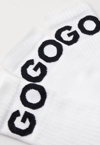 Chaussettes blanches en matériau doux avec un texte noir brodé épelant "GOGO" au niveau de la cheville. La texture paraît lisse et extensible.