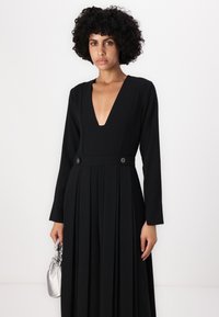 Schwarzes langärmeliges Kleid mit tiefem V-Ausschnitt, plissiertem Rock und goldfarbenen Knöpfen an der Taille. Glattes Material mit weicher Textur.