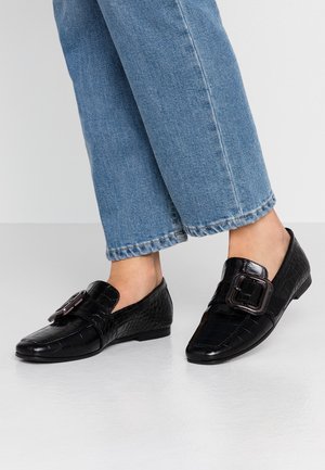 Sorte loafers med krokodillemønster og stort spænde, iført med blå cropped jeans på bar fødder mod en hvid baggrund.