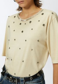 Bežina košulja kratkih rukava s metalik cvjetnim ukrasima, rebrastim izrezom i mekim teksturom, uparena s crnim remenom.
