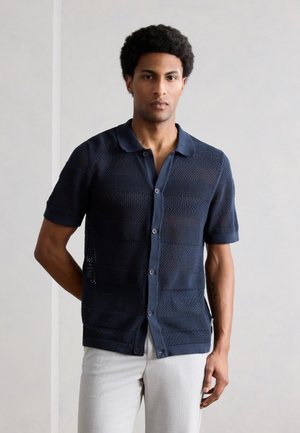 Νεαρός με κοντά σγουρά μαλλιά, ντυμένος με μπλε σκούρο κοντομάνικο πουκάμισο με κουμπιά και ελαφρώς γκρι παντελόνι, στέκεται μπροστά σε απλό φόντο.