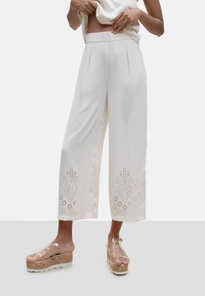 Vrouw draagt witte, hoge taille, wijde cropped broek met laser gesneden bloemenpatronen bij de zoom en transparante platformsandalen.