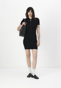 Black short-sleeve dress with a zippered collar, fitted design, and knee-length. Accessories include a black shoulder bag and black loafers.