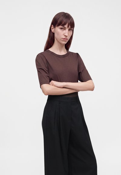 Femme aux cheveux bruns et lisses, portant un haut marron à manches courtes et un pantalon noir taille haute à larges jambes, debout les bras croisés.