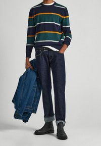 Person med randig tröja, mörka uppvikta jeans, svarta stövlar, som håller en blå denimjacka i vänster hand, höger hand i fickan.