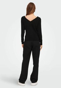 Mujer con cabello largo castaño, vistiendo un suéter negro de espalda descubierta, pantalones anchos negros y zapatillas blancas, de pie contra un fondo claro uniforme.