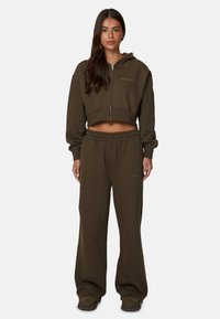 Sweat à capuche zippé vert olive, coupé, avec poches avant, associé à un pantalon de survêtement ample. Les deux pièces sont en tissu doux et texturé.