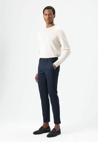 Beige gebreide trui gecombineerd met marineblauwe, slim-fit broek. Model draagt donkere loafers. Eenvoudig ontwerp en lichtgewicht stof met een gladde textuur.