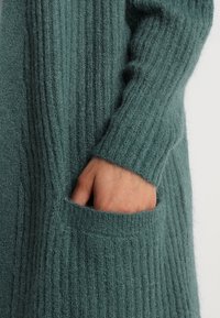 Cardigan verde in maglia con texture a coste verticali, maniche lunghe e tasca frontale, realizzato in materiale morbido e soffice.