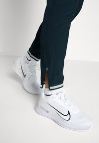 Vita atletiska skor med texturerad mesh överdel och svart swoosh, i kombination med mörka joggers med dragkedjor och randiga muddar.