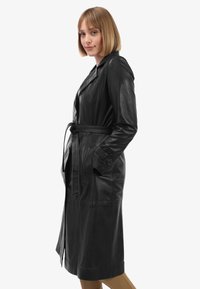 Zwarte leren trenchcoat met een tailleband, incisies op de kraag, lange mouwen en zijzakken. Glad materiaal met een aansluitend ontwerp.