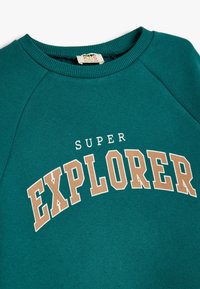 Sudadera color verde azulado con un cuello redondo acanalado. Presenta "SUPER EXPLORER" en letras grandes color beige. Tejido suave y texturizado con mangas raglán.