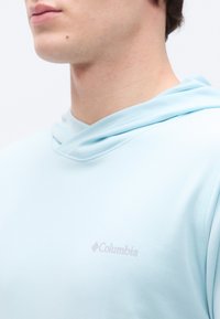 Κοντινό ενός ατόμου που φοράει ανοιχτό γαλάζιο φούτερ Columbia, εμφανίζοντας τον λαιμό, το πιγούνι και μερικό μέρος του ώμου σε απλό φόντο.