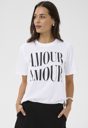 Frau trägt ein weißes T-Shirt mit schwarzem "AMOUR AMOUR" Schriftzug, schwarzen Rock, silberne Ohrringe und Armband, Hände in den Taschen, schaut nach links.