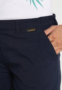 Marina stretchbyxor med elastisk midja, försedda med en UVShield-etikett och sidfickor, som visar en slät textur och skräddarsydd passform.