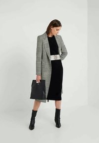 Casaco xadrez em preto e branco, usado por cima de um vestido preto com cinto decorativo. Mala de couro preta e botas até ao joelho.