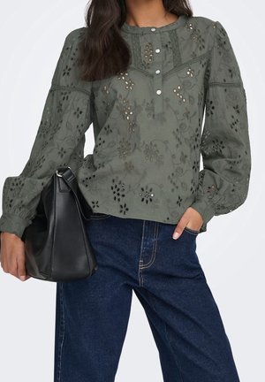 Femme portant un chemisier vert à œillets fleuris avec des manches bouffantes et un jean bleu, tenant un sac à main en cuir noir dans une main.