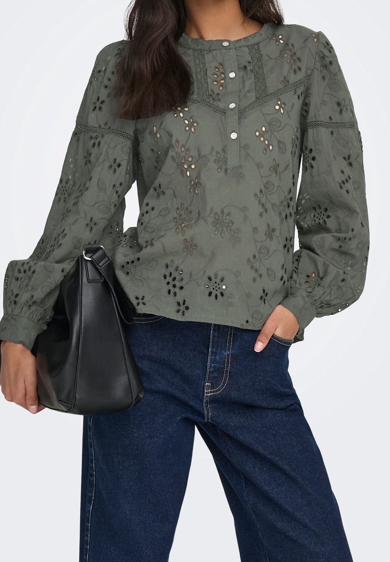 Femme portant un chemisier vert à œillets fleuris avec des manches bouffantes et un jean bleu, tenant un sac à main en cuir noir dans une main.
