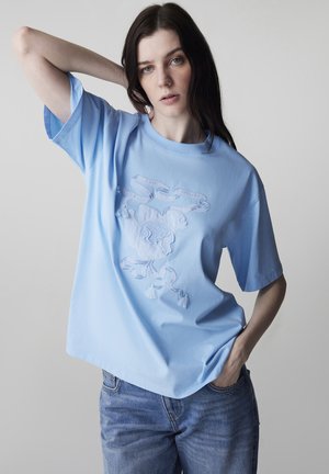 Femme aux longs cheveux bruns portant un t-shirt bleu clair avec un motif brodé d'oiseau et de ruban, assorti à un jean bleu, posant avec un bras derrière la tête.