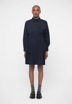 Navy blue, long-sleeve shirt dress with two chest pockets, a cinched waist belt, and a straight hem; paired with gray socks and black shoes.