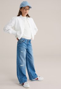 Tânăr cu mâinile pe șolduri, purtând o șapcă albastră, cămașă albă, top alb, blugi albăstri largi și pantofi sport albi cu logo roșu.