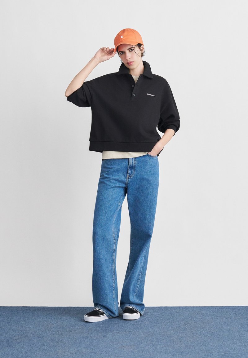 Personne portant une casquette orange, un pull noir à boutons partiels, une chemise claire en dessous, un jean bleu et des baskets noires, debout sur un tapis bleu.