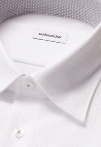 Close-up van een witte, gestructureerde overhemdkraag met twee knopen en een "seidensticker" label binnenin de kraag.