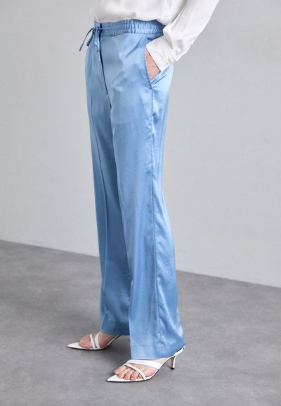 Light blue satin pants with an elastic waistband, side pockets, and a straight fit, paired with white strappy high-heeled shoes.