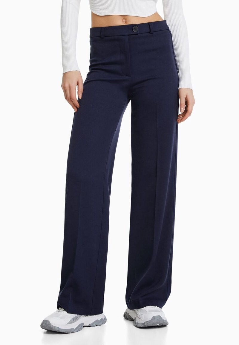 Pantalons larges marine en tissu lisse, dotés d'une fermeture à bouton et de passants de ceinture. Associés à des baskets grises pour un contraste.