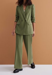 Olivově zelený propracovaný oblek s dvouřadým sakem a kalhotami širokého střihu, se hladkým povrchem a dvěma předními kapsami. Černé boty.