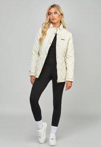 Quiltet, cremefarvet jakke med trykknapper, sidelommer og højt krave, parret med sorte leggings og hvide sneakers.