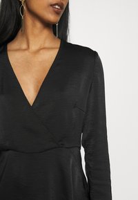 Černý wrap top s dlouhými rukávy, s hlubokým V-výstřihem, jemnou lesklou texturou a decentními stehy. Viditelné zlaté visací náušnice.