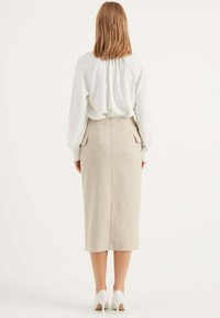Beige Bleistiftrock mit Schlitzen hinten und Seitentaschen, aus strukturiertem Stoff. Kombiniert mit einer weißen, fließenden Bluse und weißen High Heels.