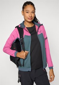 Hooded jacket, pink and teal color blocks, soft fabric, zipper details, paired with a black backpack and textured black top, functional design.
