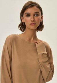 Beżowy sweter z długim rękawem, wykonany z miękkiej tkaniny, z szerokim dekoltem i luźnym krojem. Posiada mankiety na rękawach i swobodny fason. Bez wzorów ani akcentów.