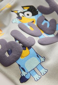 Personnage de chien de dessin animé bleu avec un visage jaune et des oreilles noires sur un tissu gris, partiellement couvert par de grandes lettres gonflées gris foncé.