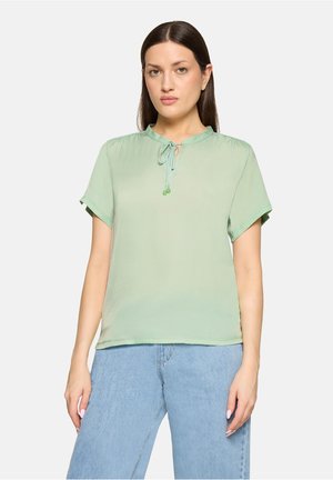 Frau trägt eine hellgrüne Kurzarmbluse mit gebundenem Ausschnitt und hellblaue Jeans mit hoher Taille, steht vor einem einfarbigen Hintergrund.