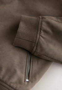 Poche zippée et poignet côtelé sur la manche d'une veste en daim marron, vue en gros plan.