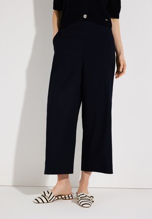Femme portant un pantalon large raccourci bleu marine et des chaussures à enfiler rayées noir et blanc avec des nœuds, debout devant un fond blanc.