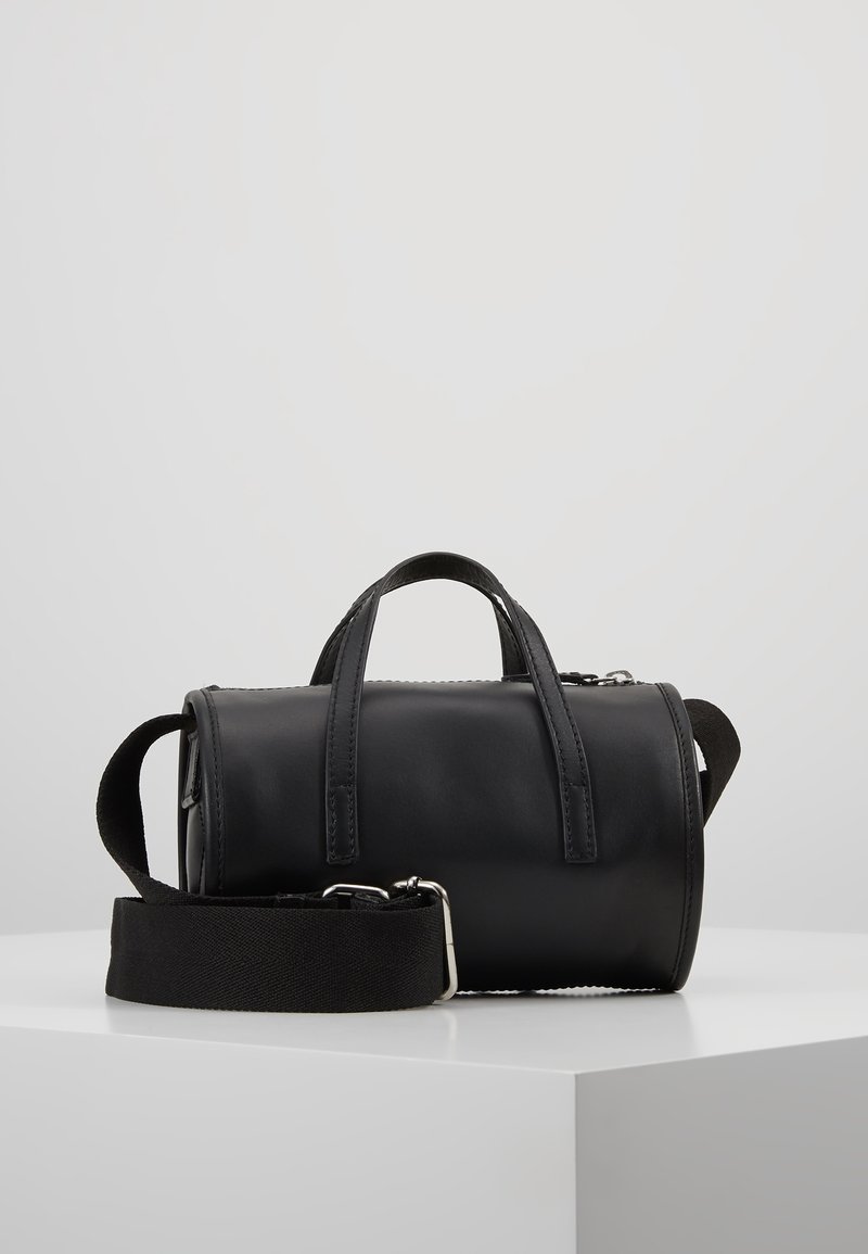 Sac cylindrique noir en cuir avec double poignées et bandoulière large en tissu détachable sur surface blanche.