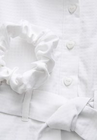 Top en tissu blanc avec un motif texturé, des boutons en forme de cœur et un chouchou assorti. Présente un détail de ceinture nouée.