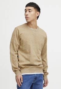 Beige gebreide trui met een V-hals, lange mouwen en ribgebreide manchetten. Gedragen over een wit shirt, gecombineerd met blauwe jeans.
