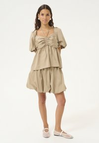 Vestito khaki con maniche a sbuffo, corpino arricciato e design a strati, con una gonna ampia e svasata. Abbinato a scarpe basse trasparenti.