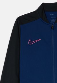 Τζάκετ Nike Dri-FIT για τρέξιμο με μπροστινό μέρος σε σκούρο μπλε, μαύρα μανίκια και ροζ κεντημένο λογότυπο· ομαλή υφή και φερμουάρ στο μπροστινό μέρος.