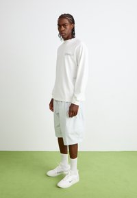 Chemise blanche à manches longues avec un logo en texte sur la poitrine, assortie à un short gris clair et des baskets blanches. Debout sur un sol vert.