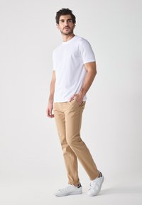 T-shirt bianca a girocollo abbinata a chinos beige e sneakers bianche. L'outfit ha una vestibilità casual con dettagli minimi e senza loghi visibili.