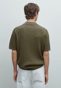 Polo shirt a maniche corte in maglia di colore verde oliva, caratterizzato da un motivo a cavo testurizzato, colletto a costine e vestibilità rilassata. Indossato con pantaloni bianchi.