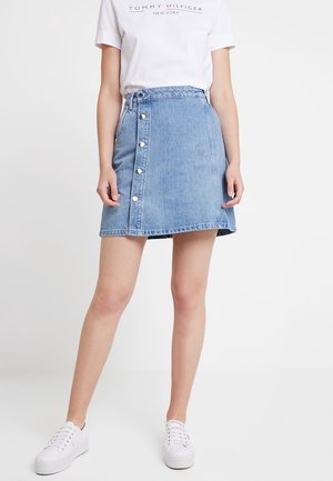 Kvinde iført hvid Tommy Hilfiger T-shirt, lyseblåt knaplukket denim mininederdel og hvide sneakers, stående mod en ensfarvet baggrund.