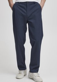 Navy blue tailored trousers with a straight fit, made of a textured fabric. Features a single button closure and side pockets. Worn with white sneakers.